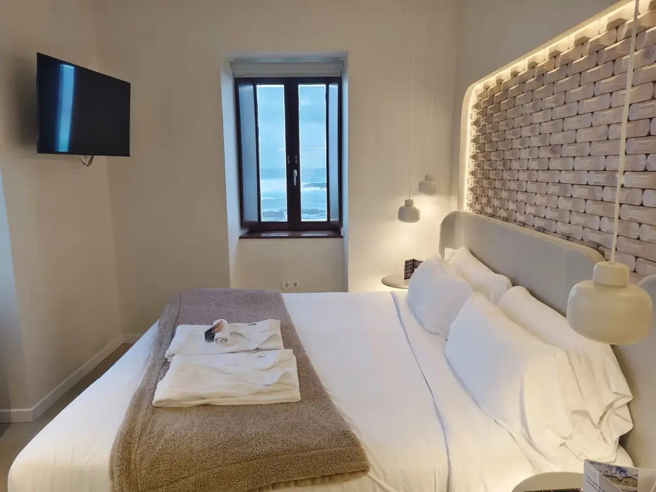Habitación de hotel con cama grande vestida de blanco, toallas dobladas sobre una manta marrón, pared de ladrillo