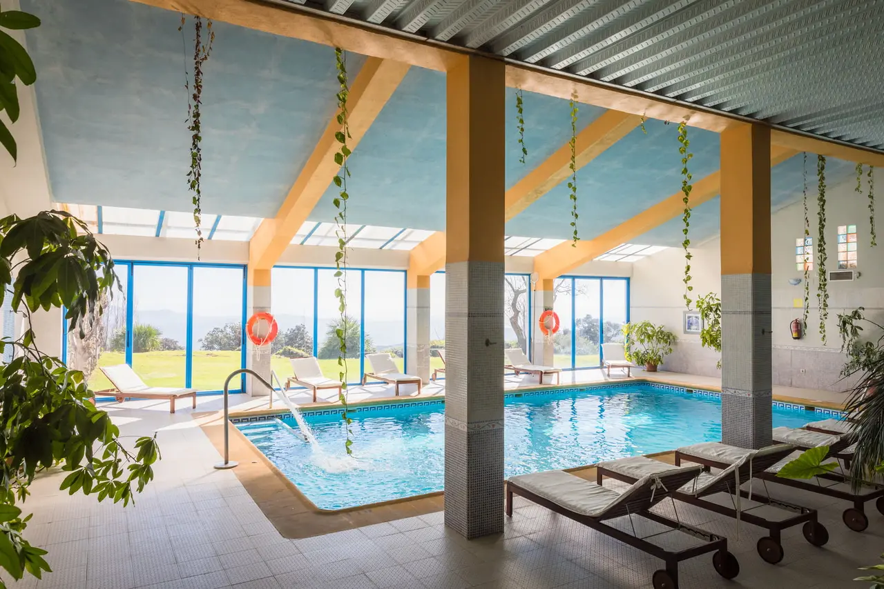 Interior de un spa privado con piscina rectangular de agua azul, chorro de agua, tumbonas de madera, pilares