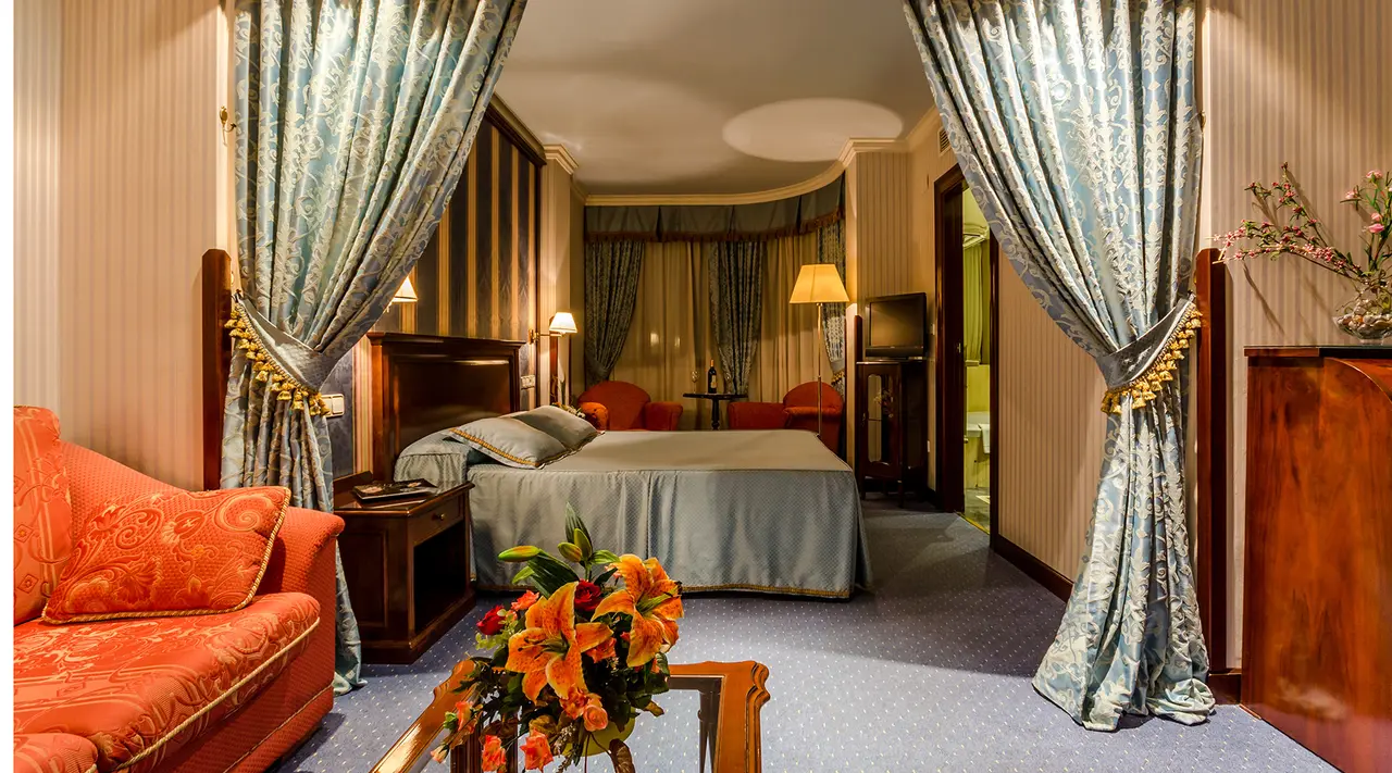 Vista de una habitación junior suite con cama grande, sofá rojo, mesa con flores, cortinas decorativas y una puerta