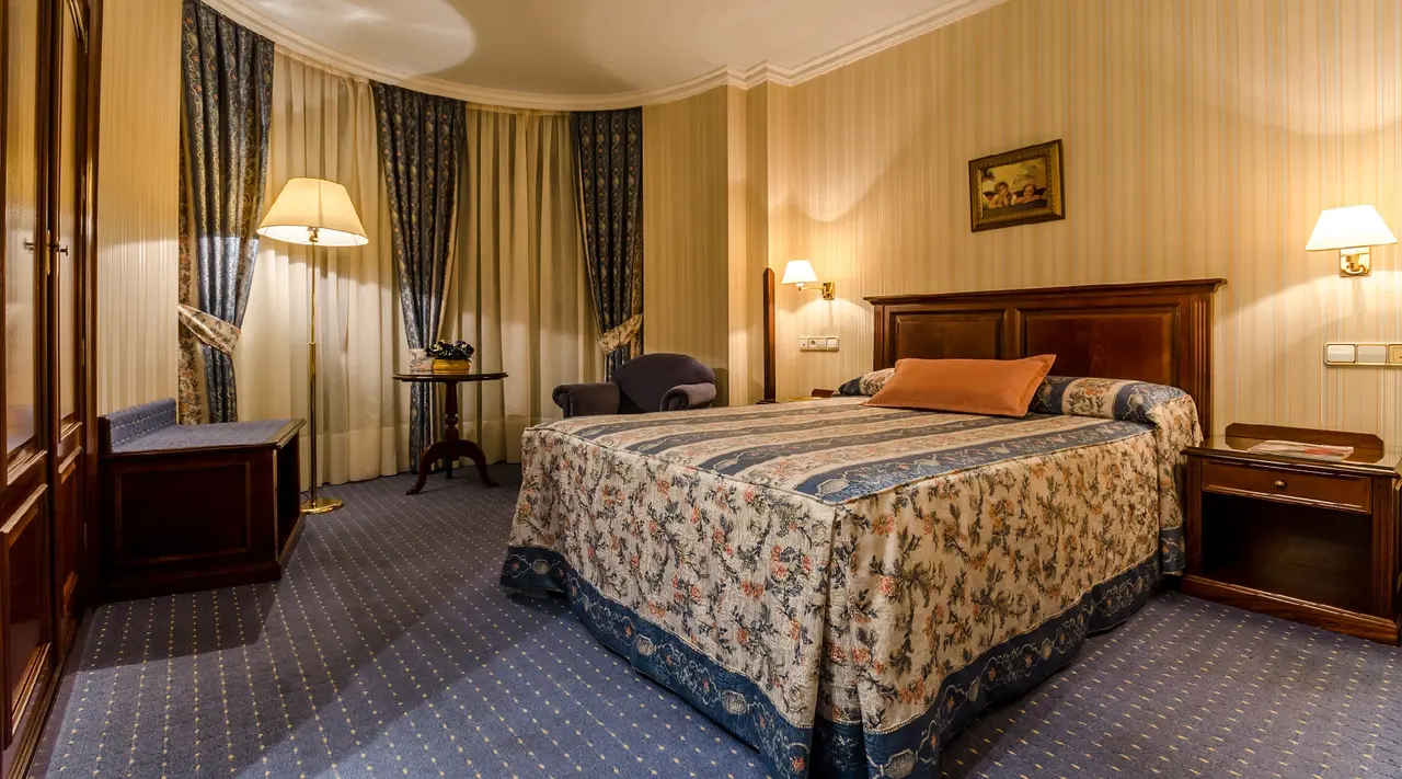 Vista de una habitación junior suite con una cama grande con colcha estampada, cabecero de madera, mesitas de noche
