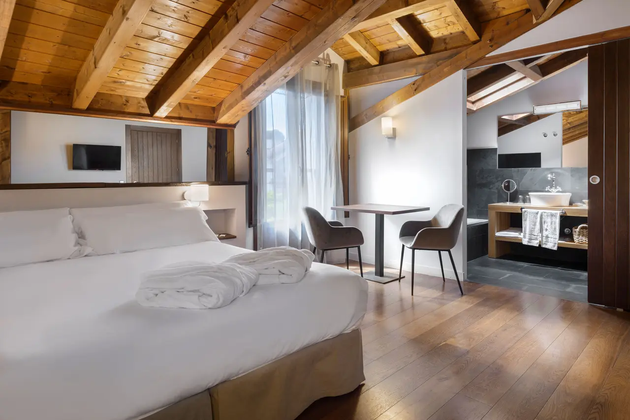 Vista de una suite con cama grande vestida de blanco, vigas de madera en el techo, zona de estar con mesa y sillas