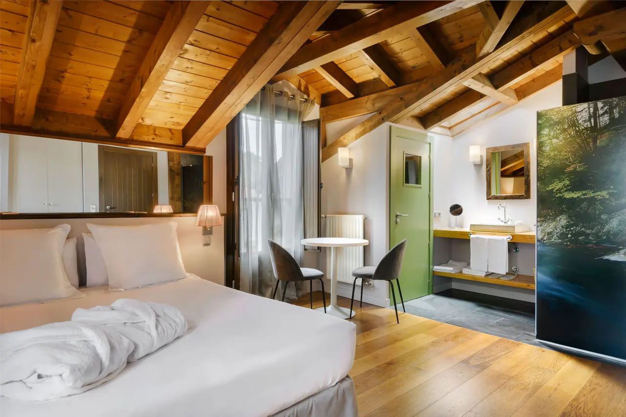 Vista de una suite con cama doble grande con sábanas blancas, techo abuhardillado de madera con vigas vistas, zona