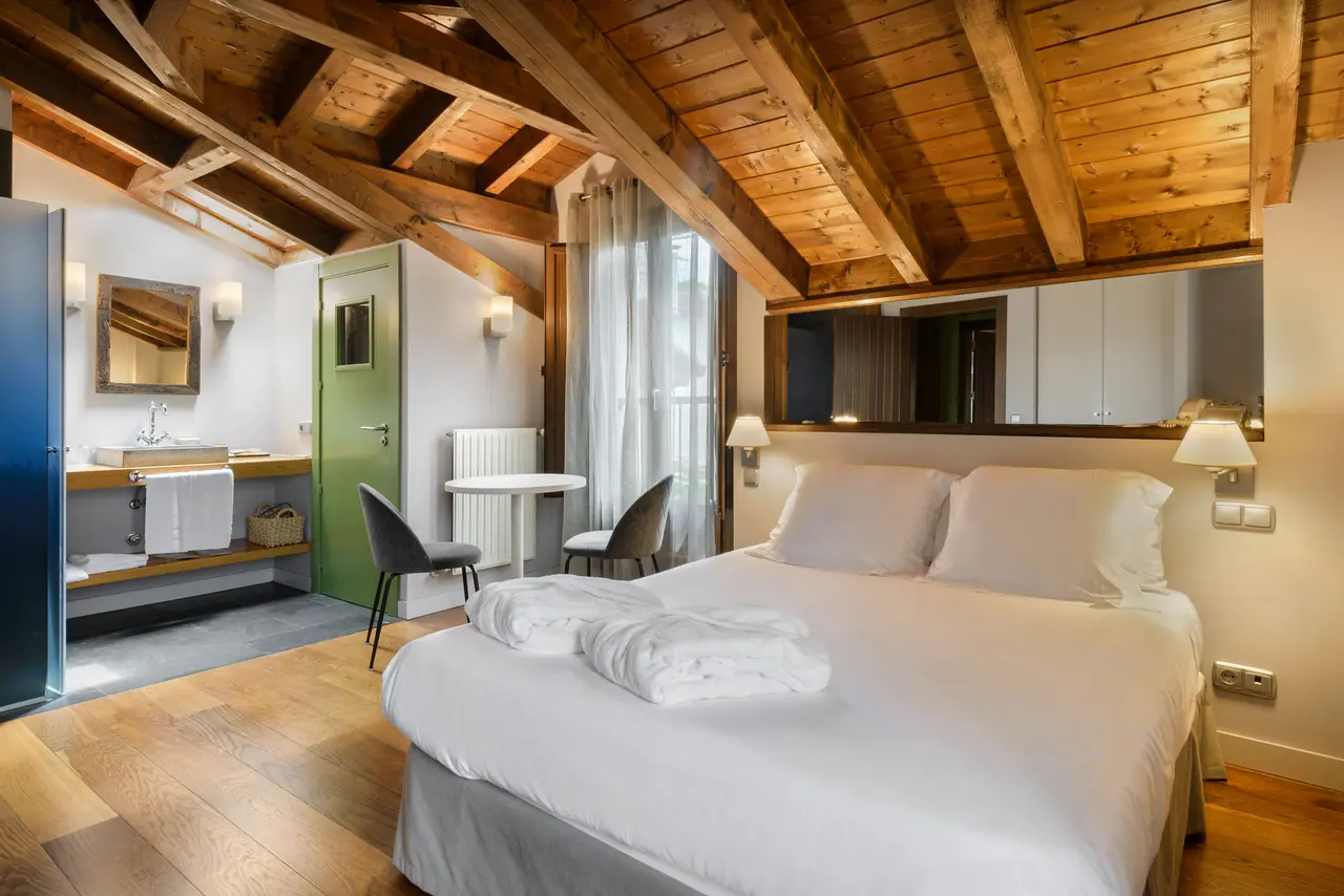 Vista de una habitación de hotel con techo abuhardillado de vigas de madera, una cama grande con sábanas blancas