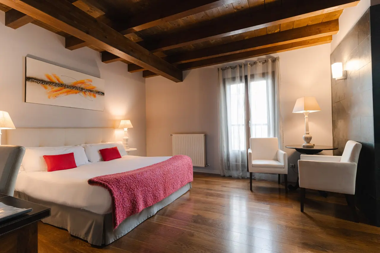 Habitación de hotel con cama doble grande vestida con edredón blanco y manta roja, dos sillones blancos