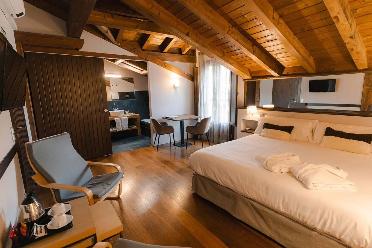 Vista de una suite con techo abuhardillado de vigas de madera, cama grande con ropa blanca, sillón azul, mesa