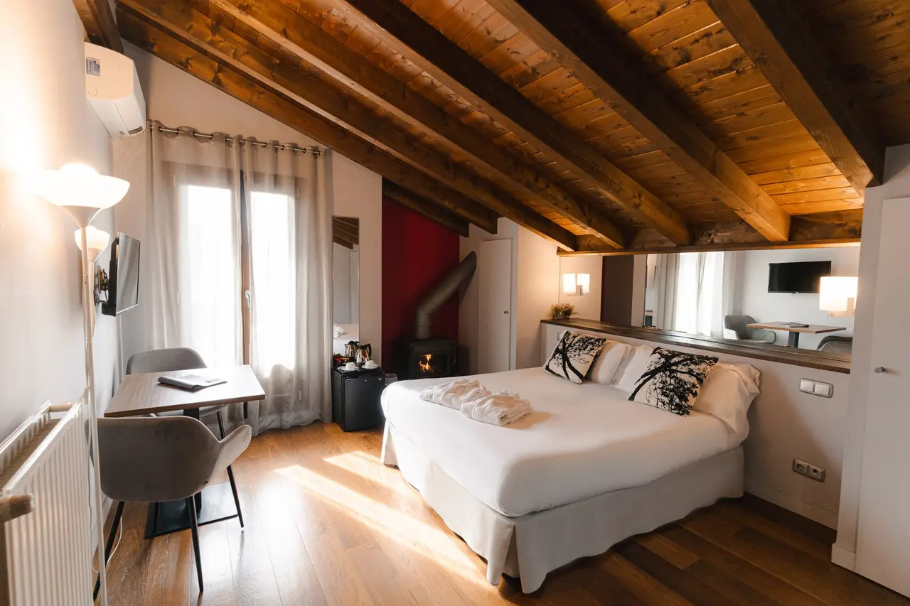 Vista de una suite con cama doble grande vestida de blanco, vigas de madera en el techo inclinado, chimenea de leña