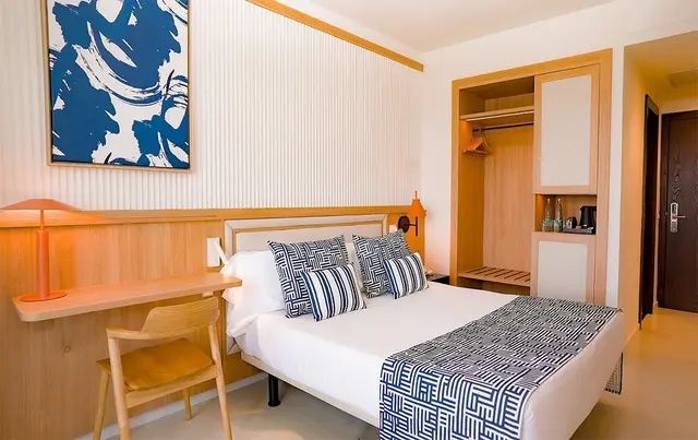 Vista de una junior suite con cama doble, escritorio de madera con silla, lámpara de mesa, armario abierto con balda