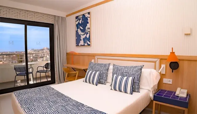 Vista de una junior suite con cama doble, cabecero de madera, almohadas y cojines de rayas azules y blancas, mesita