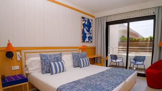 Vista de una junior suite con cama doble, cabecero de madera, cojines azules y blancos, mesita de noche, sillón rojo