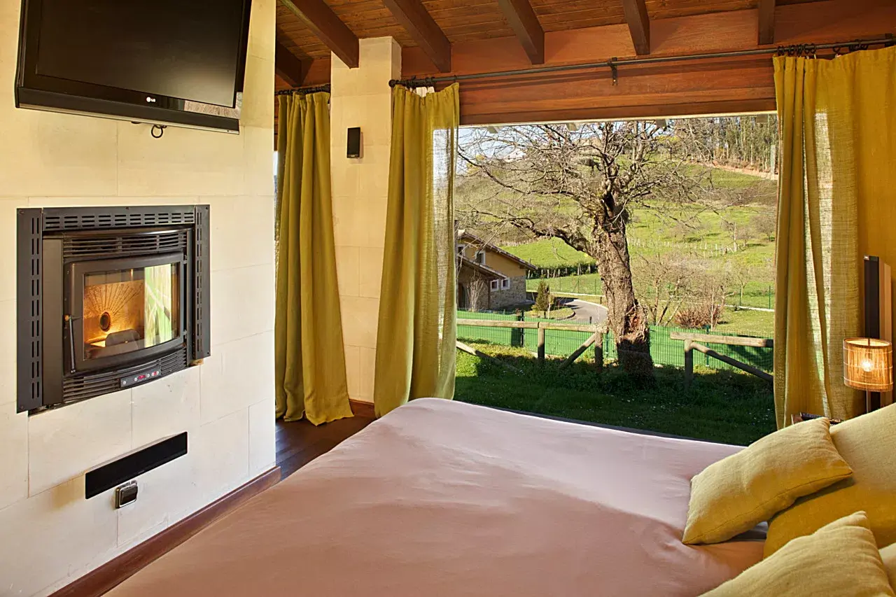 Interior de una habitación con cama grande, chimenea empotrada en la pared, televisor montado, cortinas amarillas