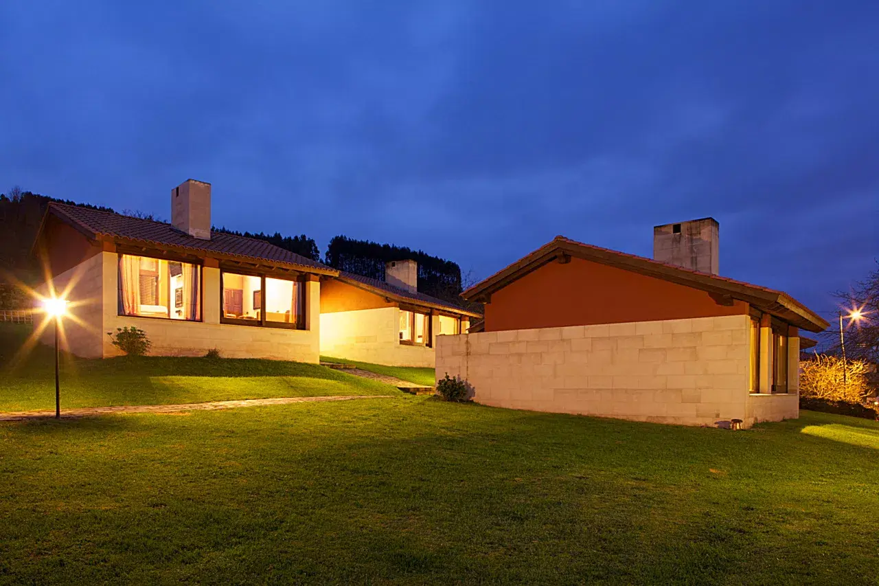 Vista exterior de una casa rural de una planta con fachada blanca y tejado a dos aguas, iluminada al atardecer.