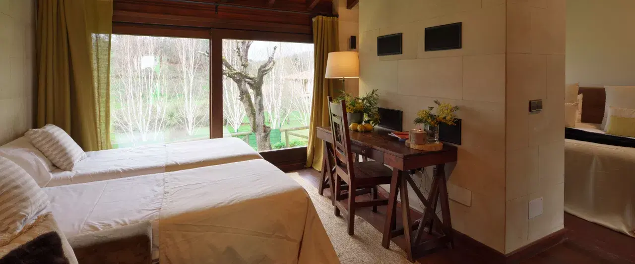 Habitación de hotel con dos camas individuales, escritorio de madera con silla, lámpara y flores, y una gran ventana