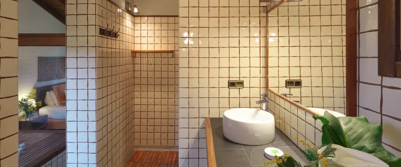Baño con ducha de pared, lavabo redondo blanco sobre encimera de piedra, espejo y azulejos beige en las paredes.