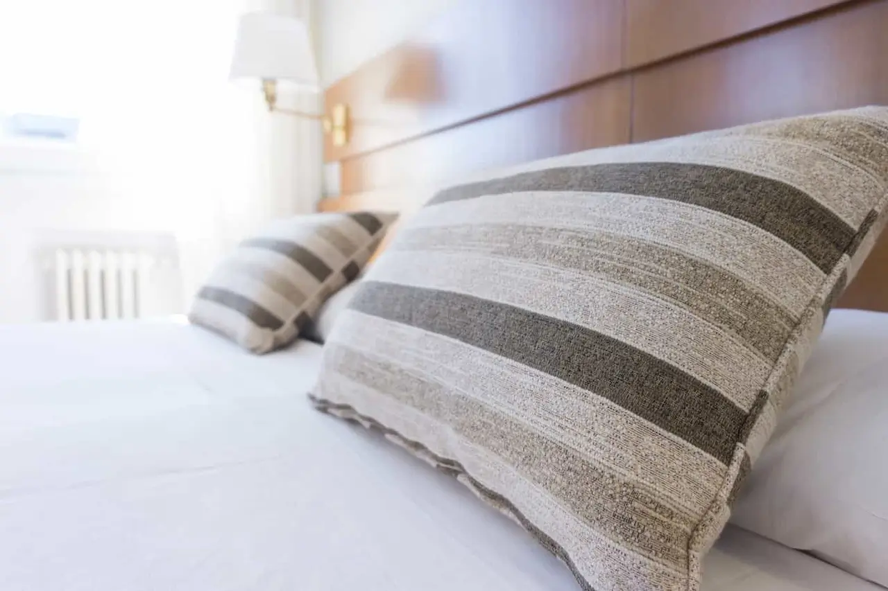 Cama de habitación con almohadas a rayas y cabecero de madera, junto a una lámpara de pared.