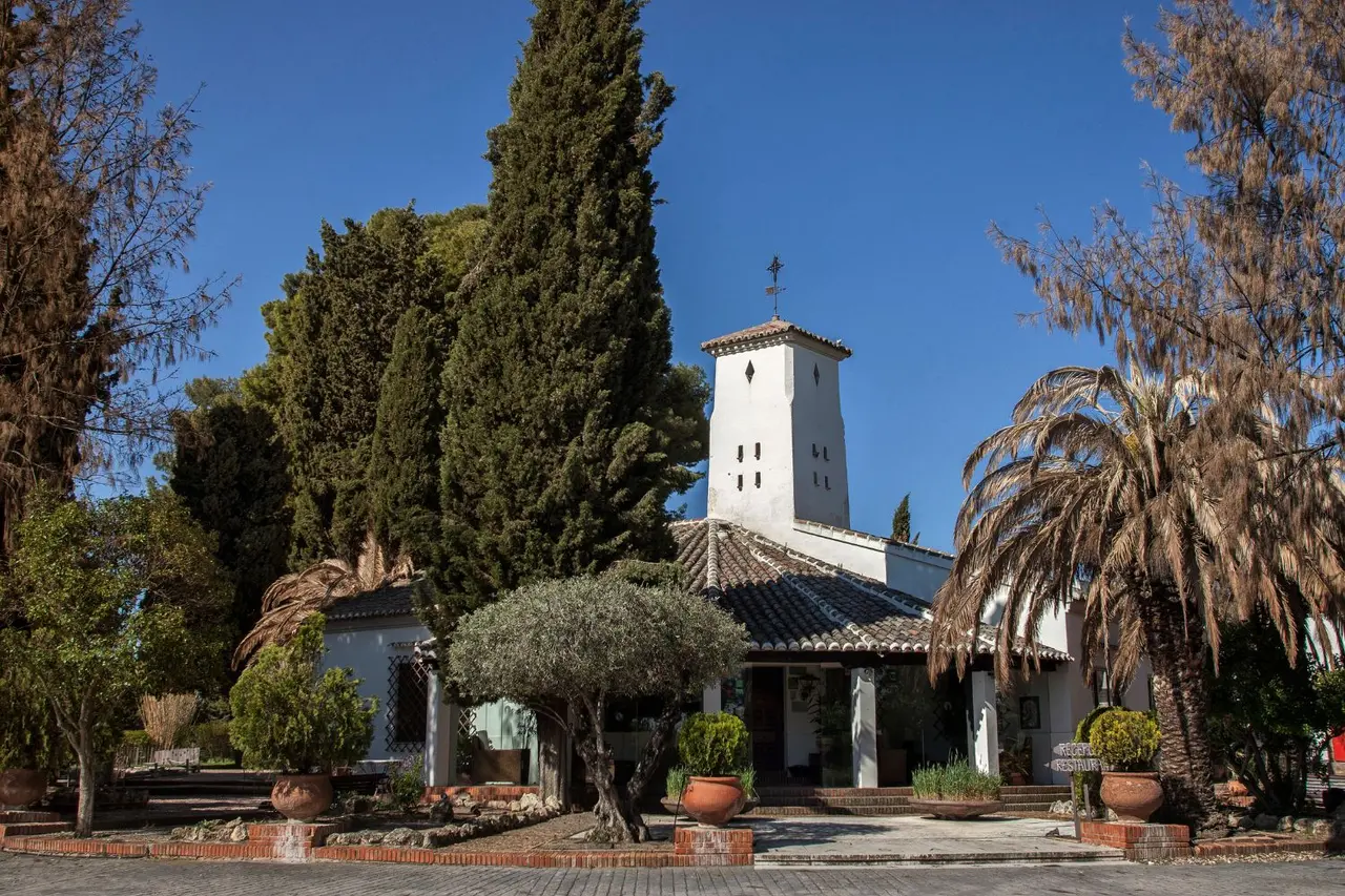 Vista exterior de un edificio blanco con torre y tejado de teja, rodeado de jardines con árboles, palmeras, arbustos