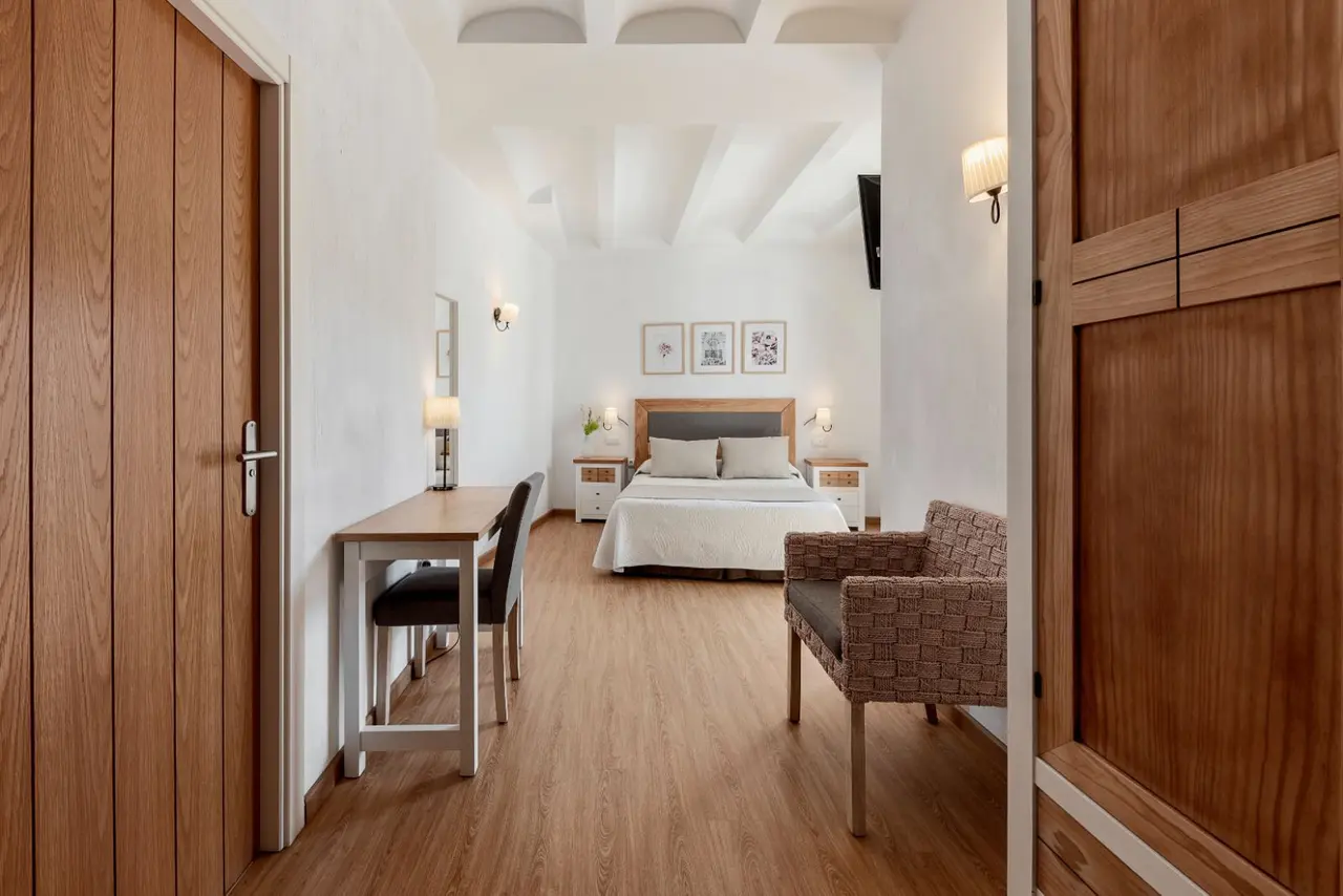 Vista de una habitación de hotel con cama doble al fondo, escritorio con silla a la izquierda, sillón de mimbre