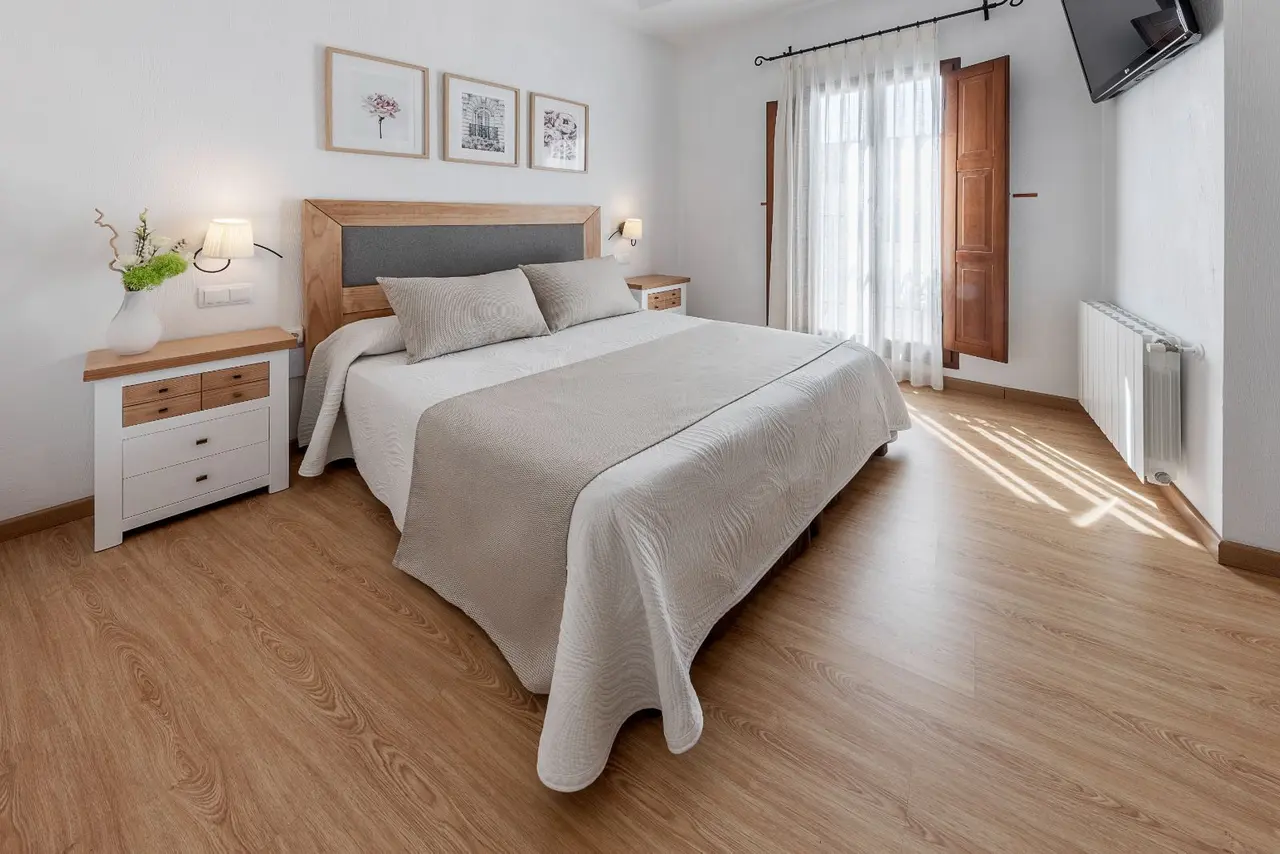 Habitación con cama doble, cabecero de madera, mesitas de noche blancas, suelo de madera clara, ventana con cortinas
