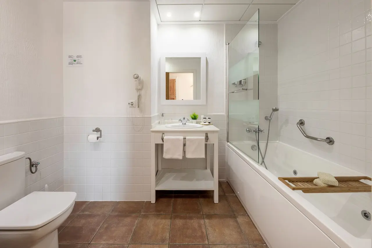 Baño completo con paredes y suelos de azulejos blancos y marrones, inodoro blanco, lavabo con espejo, toallas blancas