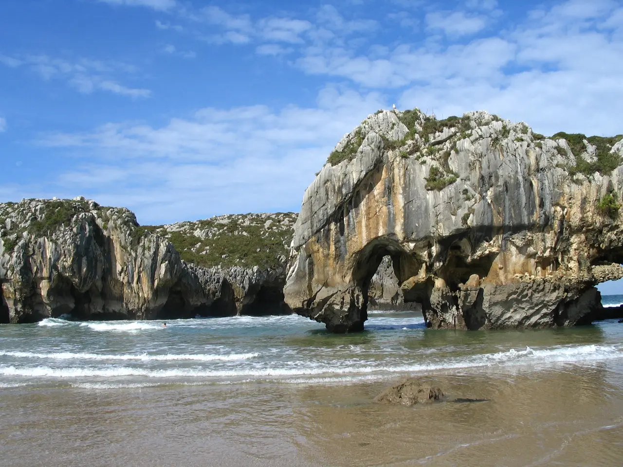 Vista de un arco natural de roca en un acantilado costero, con olas rompiendo en la orilla arenosa y un cielo azul