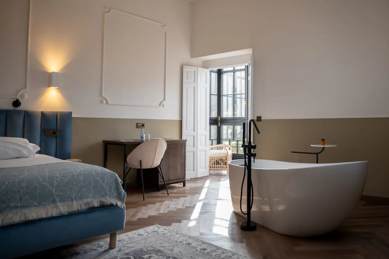 Habitación con cama azul a la izquierda, escritorio con silla, y bañera blanca independiente con grifería negra