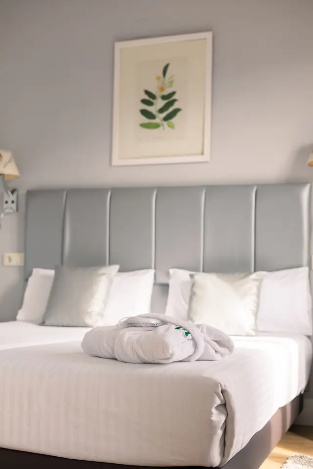 Cama doble en una habitación de hotel con cabecero gris acolchado, múltiples almohadas blancas, un albornoz blanco