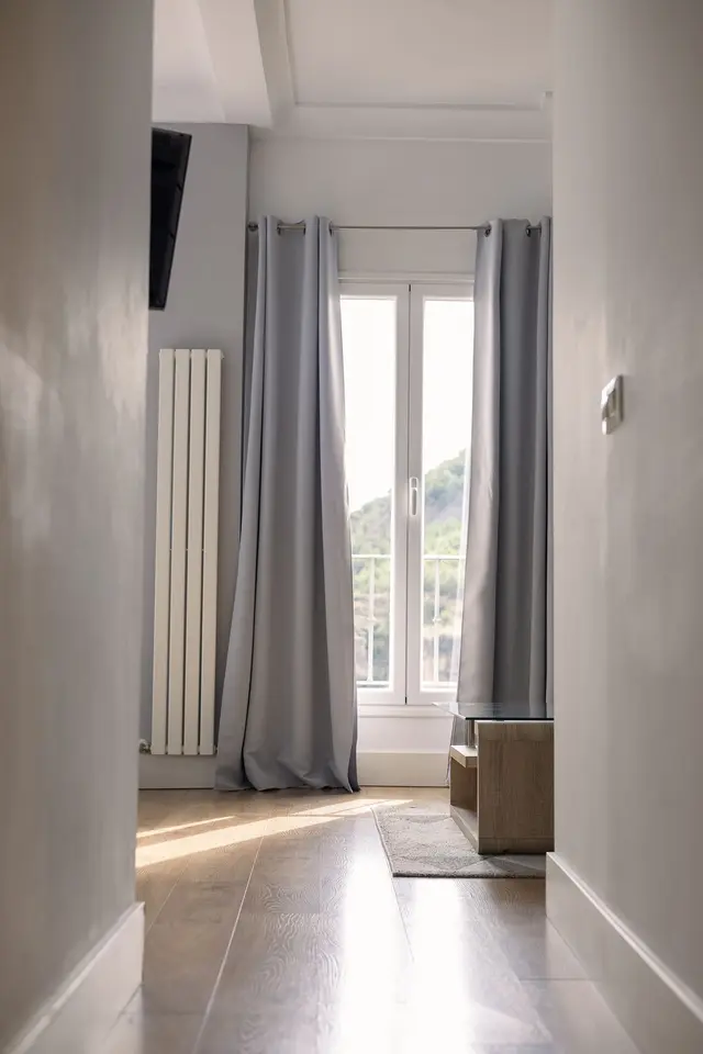 Vista de una habitación con suelo de madera clara, una ventana grande con cortinas grises que deja entrar la luz