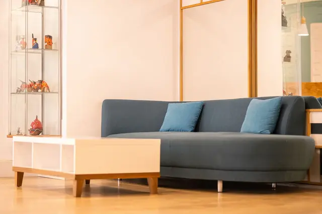 Interior de una habitación con un sofá azul curvado, dos cojines azules, una mesa de centro blanca con patas de madera