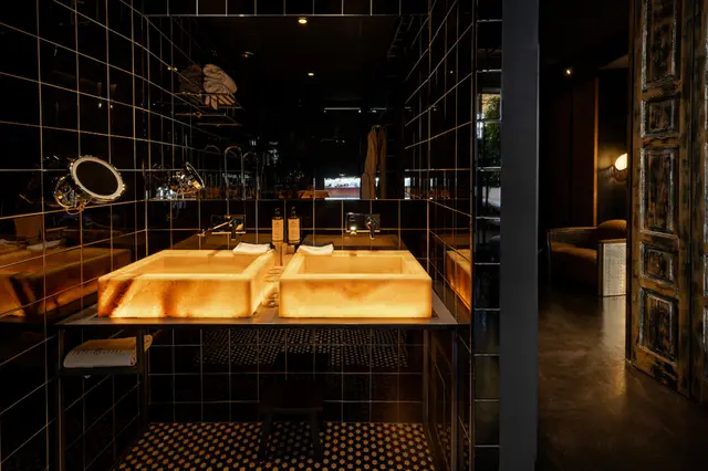Doble lavabo de piedra iluminado en baño con paredes de azulejos negros y suelo de mosaico.