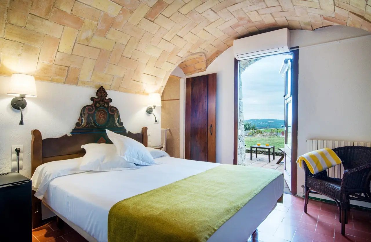 Habitación con cama doble, cabecero de madera tallada, techo abovedado de piedra, suelo de terracota y puerta abierta