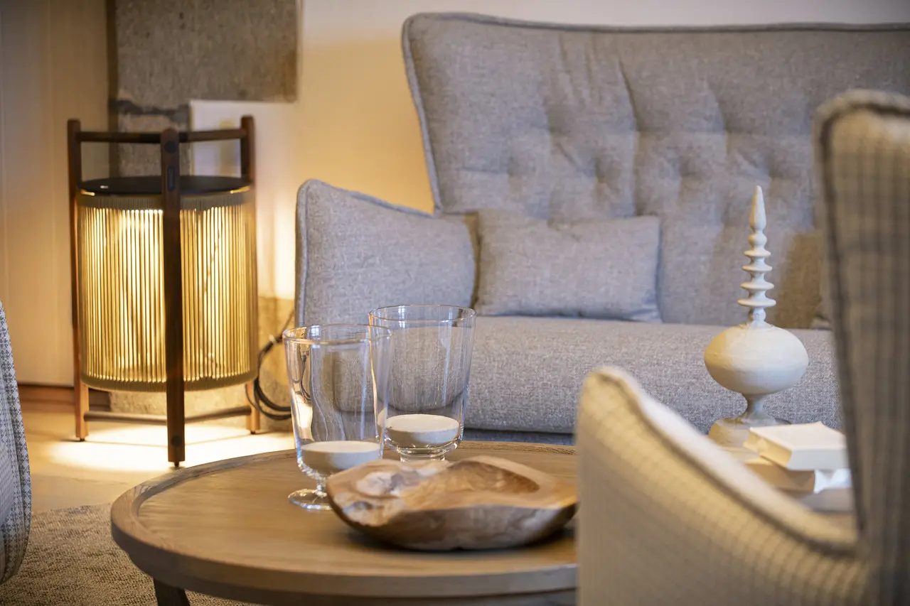 Interior de sala de relax con sofá gris, mesa de centro redonda de madera con vasos, vela y cuenco decorativo, lámpara