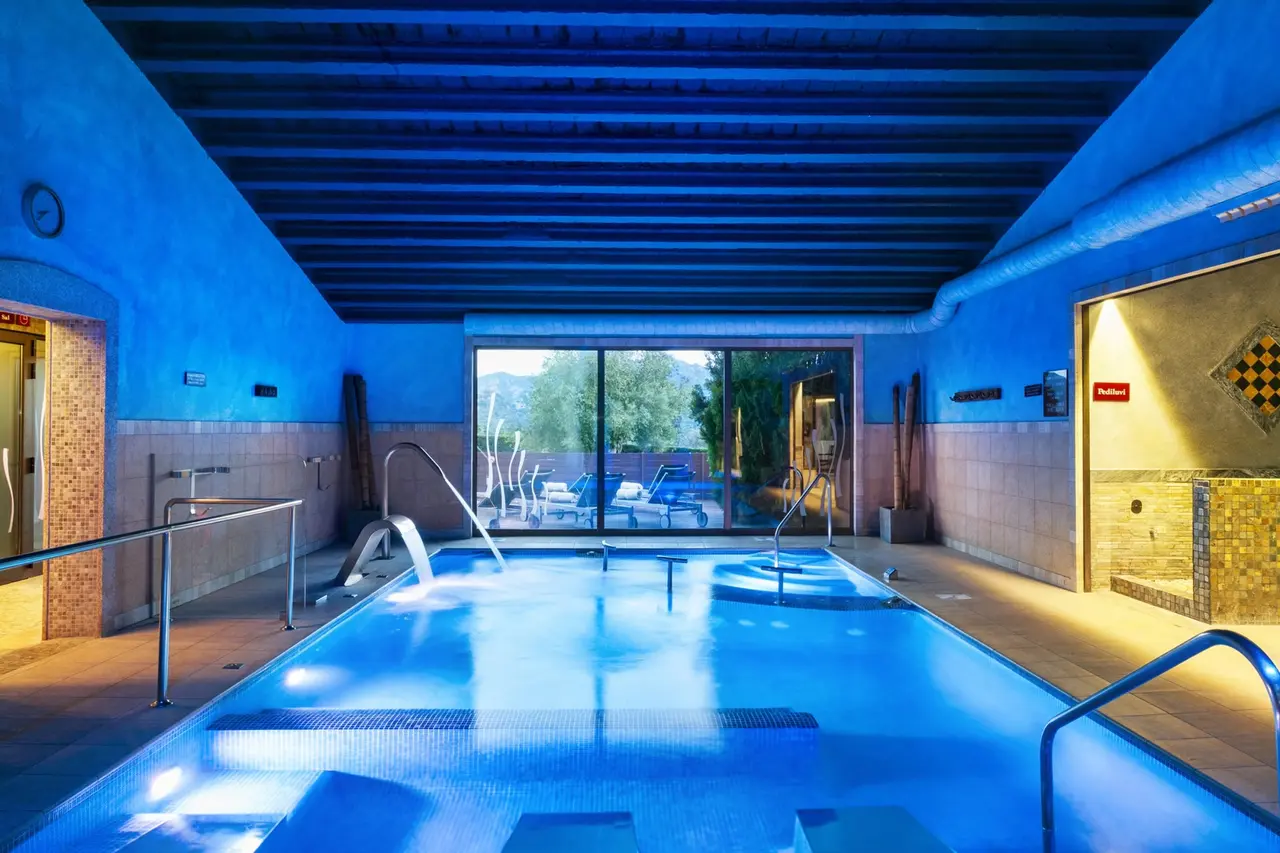 Interior de un spa nocturno con piscina iluminada en tonos azules, bancos sumergidos, barandillas metálicas y grandes