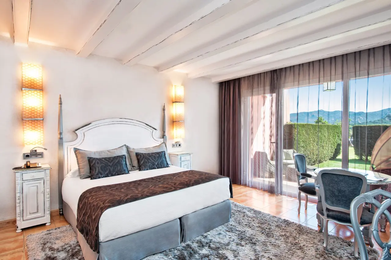Habitación con cama doble, cabecero blanco, mesitas de noche, lámparas de pared, alfombra, sillas y mesa