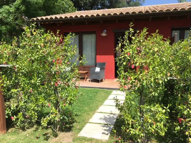 Vista exterior de una casa con fachada roja y tejado de teja, rodeada de vegetación y arbustos con flores.