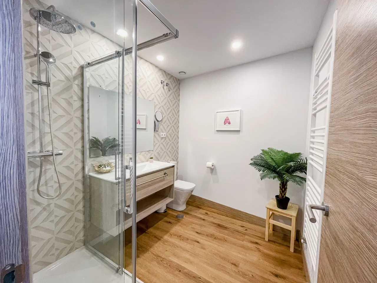 Baño moderno con ducha de cristal, lavabo de madera, inodoro, toallero eléctrico y planta en un taburete de madera.