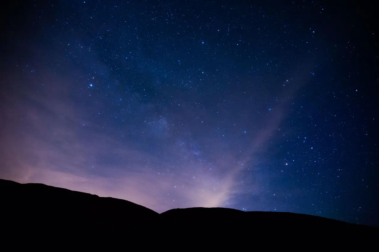 Cielo nocturno estrellado con la Vía Láctea visible sobre el horizonte oscuro de una colina.