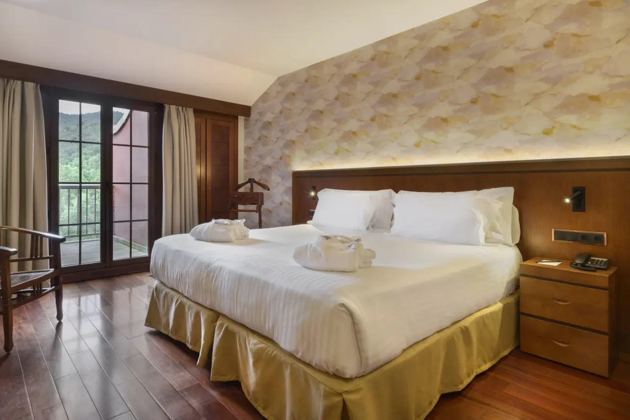 Habitación de hotel con cama doble grande vestida de blanco, cojines decorativos en forma de cisne, cabecero de madera