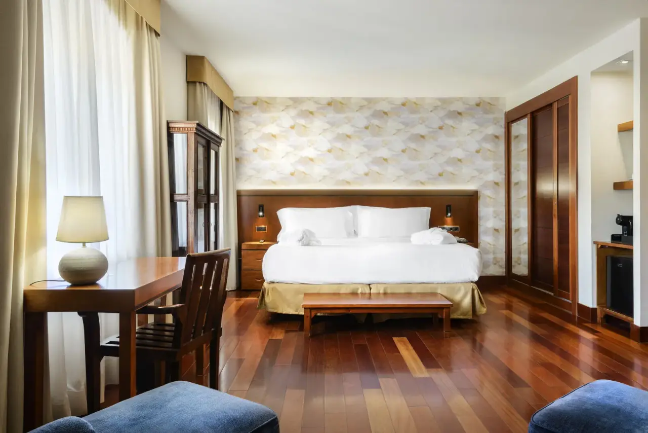 Habitación de hotel con cama doble grande vestida de blanco, cabecero de madera, mesitas de noche, escritorio