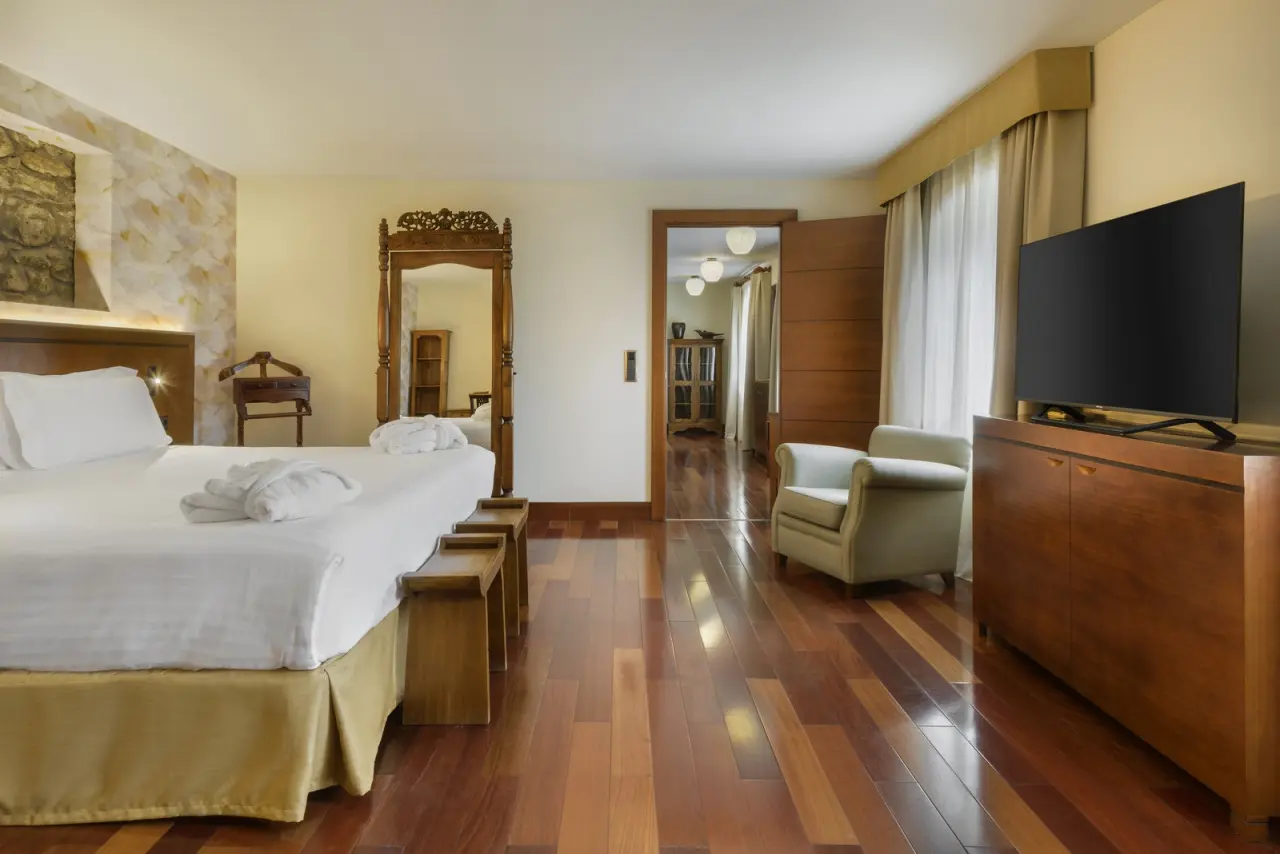 Habitación de hotel con cama doble grande vestida de blanco, dos banquetas de madera al pie, sillón beige