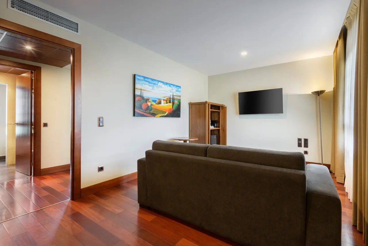 Sala de estar con sofá marrón, suelo de madera, televisor en la pared, cuadro colorido, armario de madera y puerta