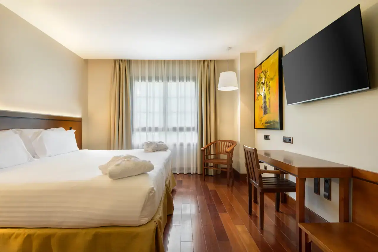 Habitación de hotel con cama doble grande vestida de blanco, toallas enrolladas, escritorio de madera con silla