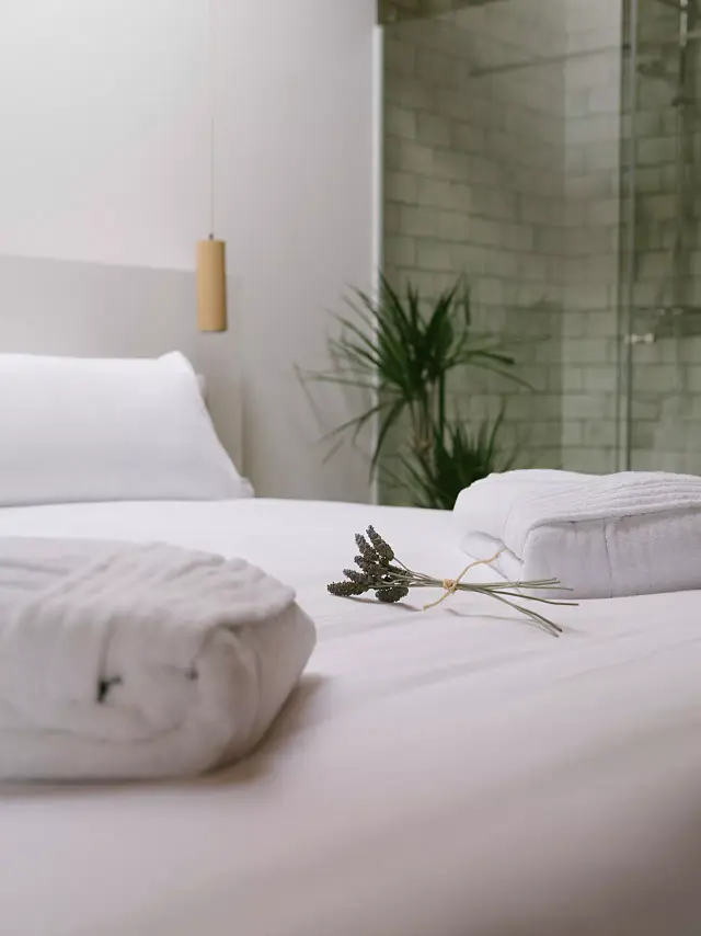 Vista de una habitación de hotel con una cama grande vestida de blanco, toallas enrolladas y un ramo de lavanda sobre la