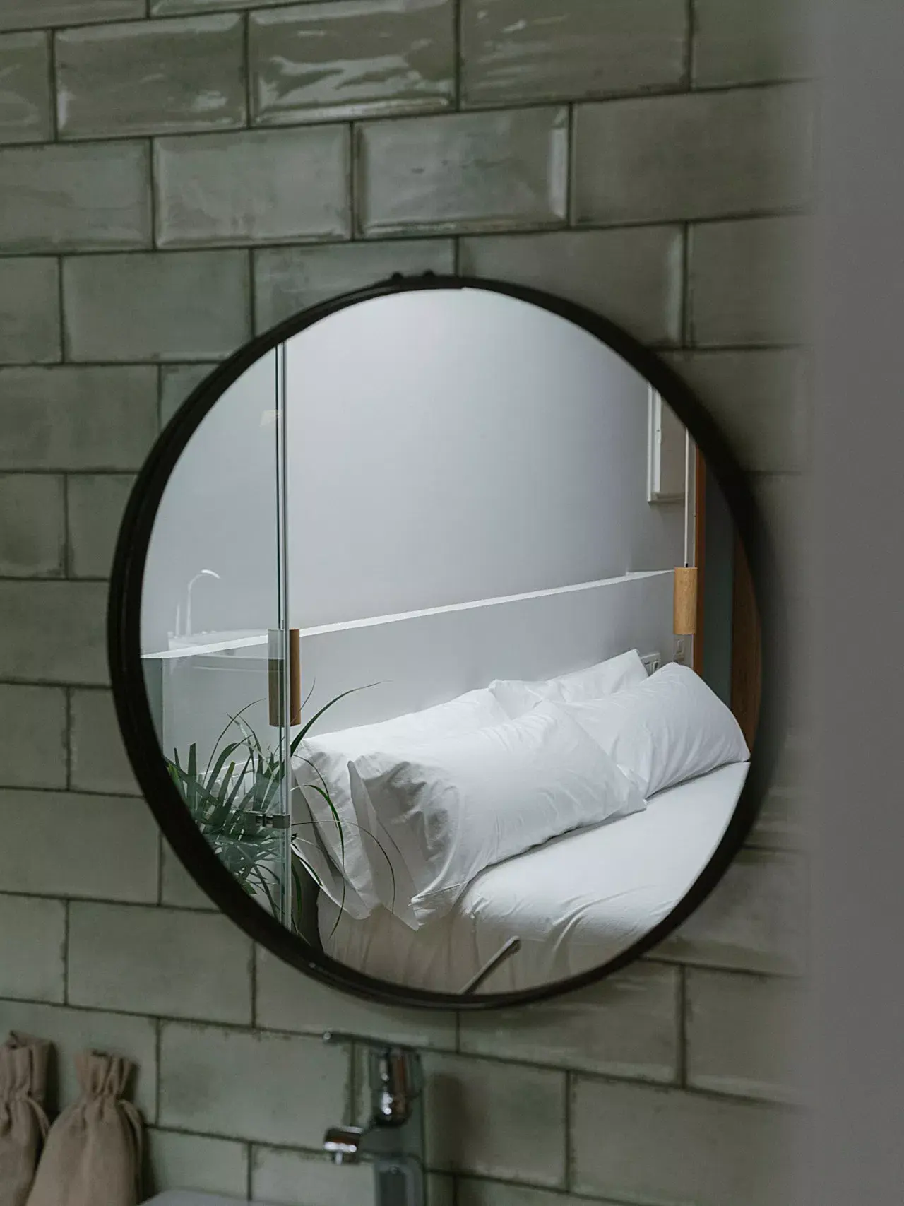 Espejo circular con marco negro montado en una pared de azulejos grises, reflejando una cama con sábanas y almohadas