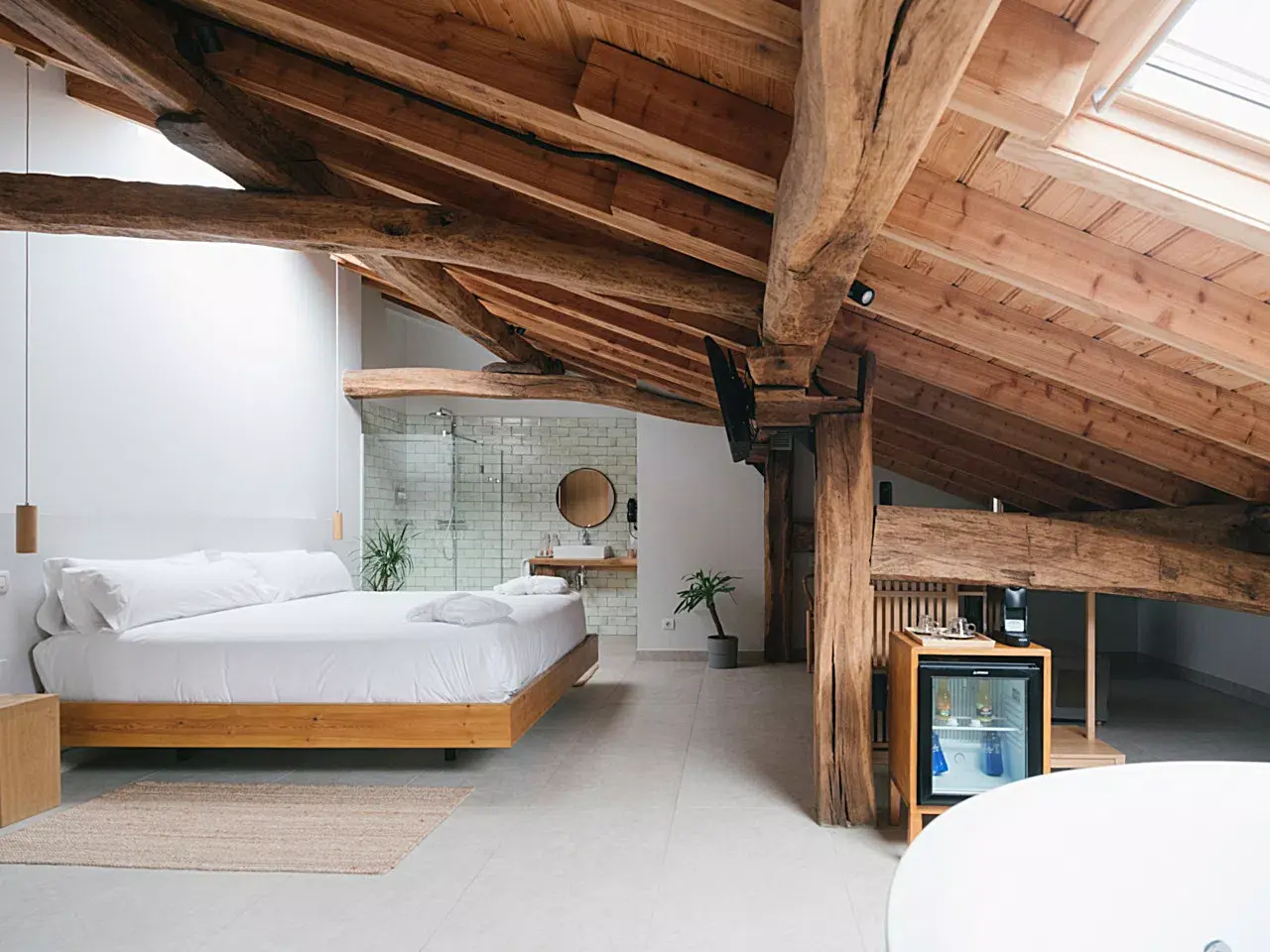 Vista de una habitación suite con techo abuhardillado de vigas de madera vistas, cama grande con base de madera y ropa