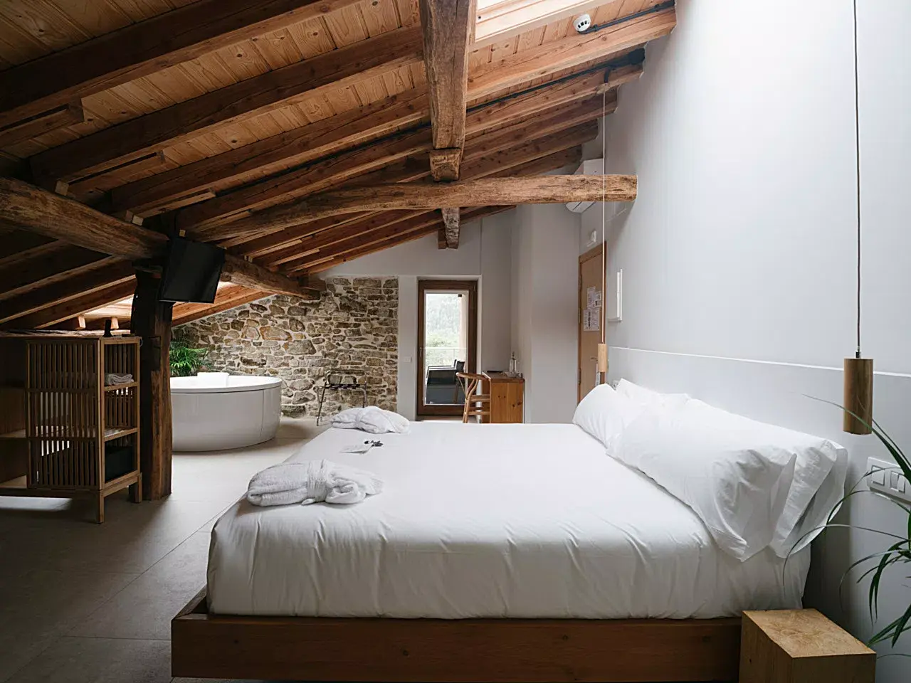 Habitación suite con cama grande vestida de blanco, vigas de madera en el techo, bañera de hidromasaje redonda