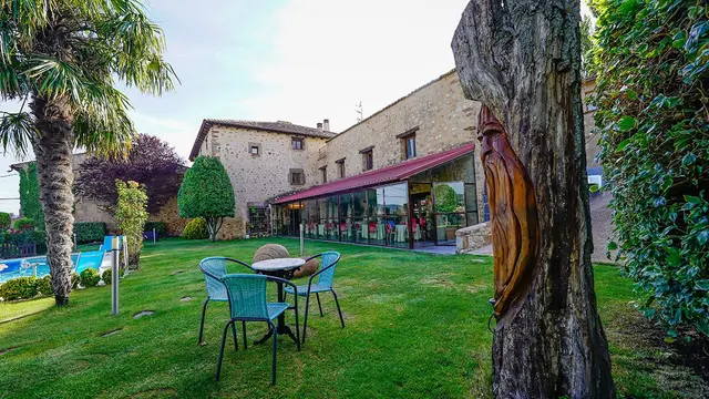Vista exterior del jardín del hotel con césped, mesas y sillas, una piscina al fondo, árboles y el edificio de piedra