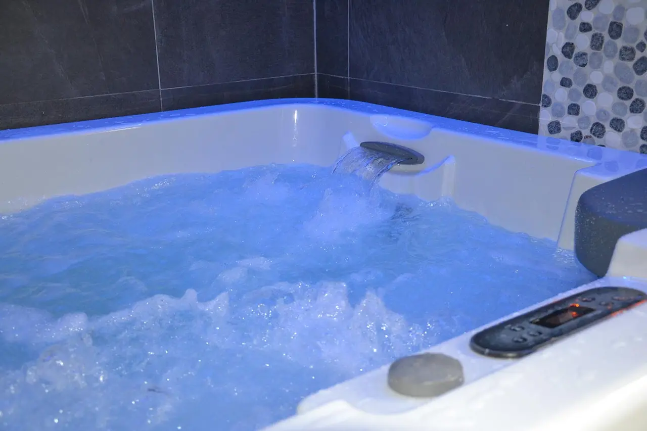 Jacuzzi blanco con agua burbujeante iluminada en azul y cascada en funcionamiento, rodeado de azulejos oscuros.
