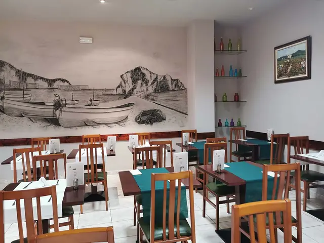 Interior de un restaurante con mesas y sillas de madera, paredes blancas y un gran mural en blanco y negro