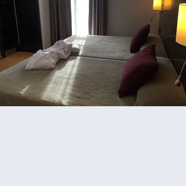 Habitación de hotel con dos camas grandes, almohadas granates, batas blancas dobladas y luz natural entrando