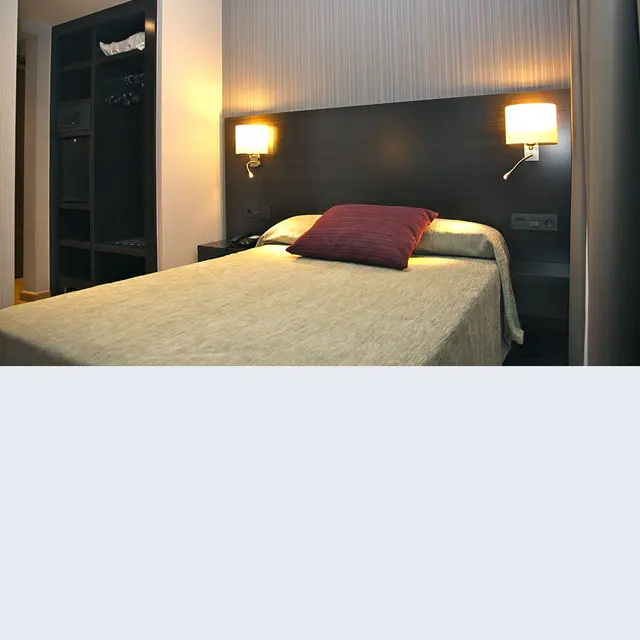 Habitación de hotel con cama grande, cabecero oscuro, dos lámparas de pared encendidas y armario abierto al lado.