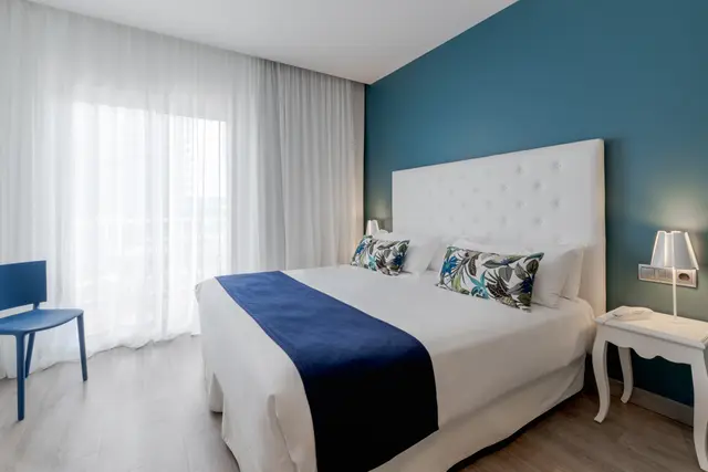 Habitación de suite con cama doble grande vestida de blanco con una franja azul, cabecero blanco acolchado, mesitas